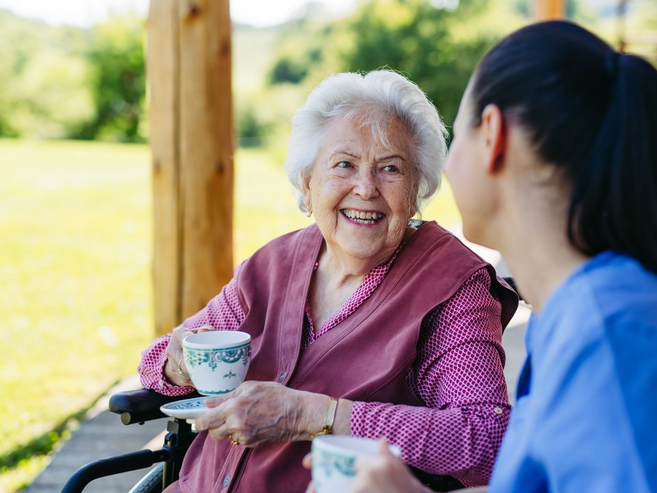 Female caregiver spending time with elderly woman, drinking coffee and talking. Nurse and senior woman in wheelchair enjoying a warm day outdoors.