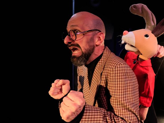 Bühnendarsteller mit Hasenpuppe, der Mann mit Brille und Bart schreit, Fäuste geballt.